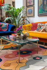 A vibrant living room featuring a Cosmic Tides hand-tufted area rug with abstract patterns in pink, yellow, and blue hues. The wool rug enhances the space, complemented by a blue sofa and a yellow couch adorned with colorful cushions. A glass coffee table with decorative items sits atop the large area rug, perfect for adding style to any living room area.