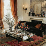 A beautifully decorated living room featuring the Countryside Charm Hand Knotted Area Rug. This vintage wool square rug adds a touch of elegance to the space, with its intricate patterns and warm colors. The rug perfectly complements the sophisticated setting with a black velvet sofa and a unique glass coffee table adorned with books and floral arrangements. Ideal for an office or living room, this square area rug enhances the cozy atmosphere.