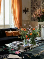 Elegant living room featuring the Crane & Pink Petal Whimsy Hand Knotted Area Rug, showcasing intricate crane and floral designs. This 8x10 Persian-inspired square rug adds a touch of whimsy and sophistication to the space. The wool rug complements the room's decor with its artistic detail, making it perfect for area rugs for living room settings. The cozy ambiance is enhanced by the warm tones of the orange curtains and floral arrangements.