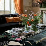 Elegant living room featuring a Crane & Pink Petal Whimsy Hand Knotted Area Rug. This Persian-inspired wool rug showcases a charming design with cranes and pink petals, perfect for adding a touch of whimsy to your living space. Ideal as a square rug or an 8x10 living room centerpiece, it complements the cozy ambiance with a black sofa adorned with orange cushions and a stylish glass coffee table.