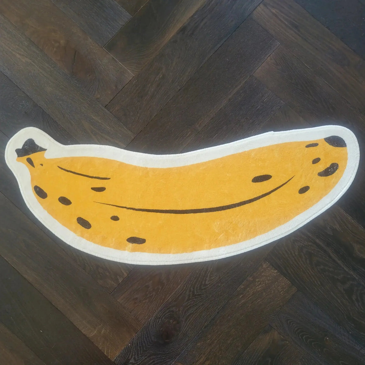 A fun and quirky area rug shaped like a banana, featuring a bright yellow color with black spots, lying on a dark wooden floor. Perfect for adding a playful touch to any living space.