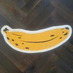A fun and quirky area rug shaped like a banana, featuring a bright yellow color with black spots, lying on a dark wooden floor. Perfect for adding a playful touch to any living space.