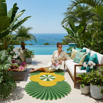 Woman sitting on a patio with ocean view, surrounded by plants and a colorful rug.