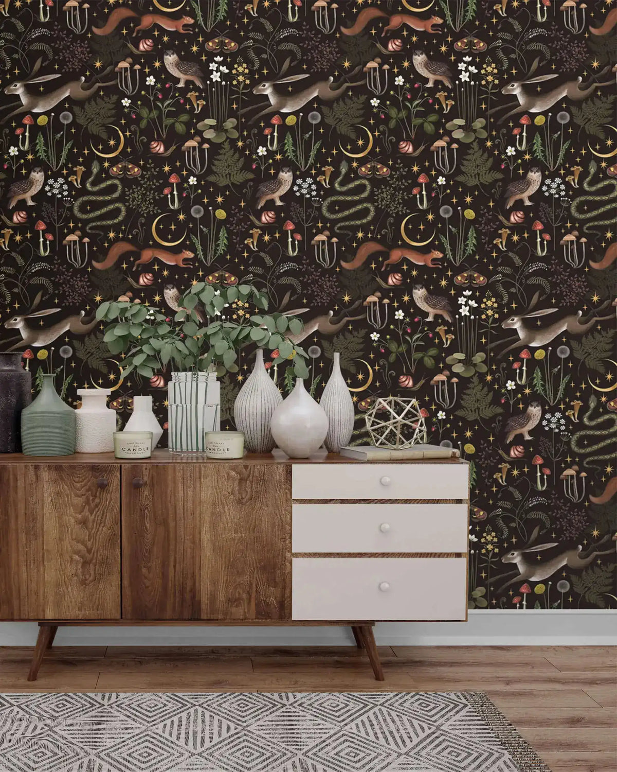 Enchanted Wilderness wallpaper behind mid-century sideboard with ceramic vases and greenery; dark patterned wall, wood floor
