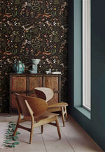 Enchanted Wilderness wallpaper behind wooden lounge chair and antique cabinet; dark pattern with forest creatures and botanicals