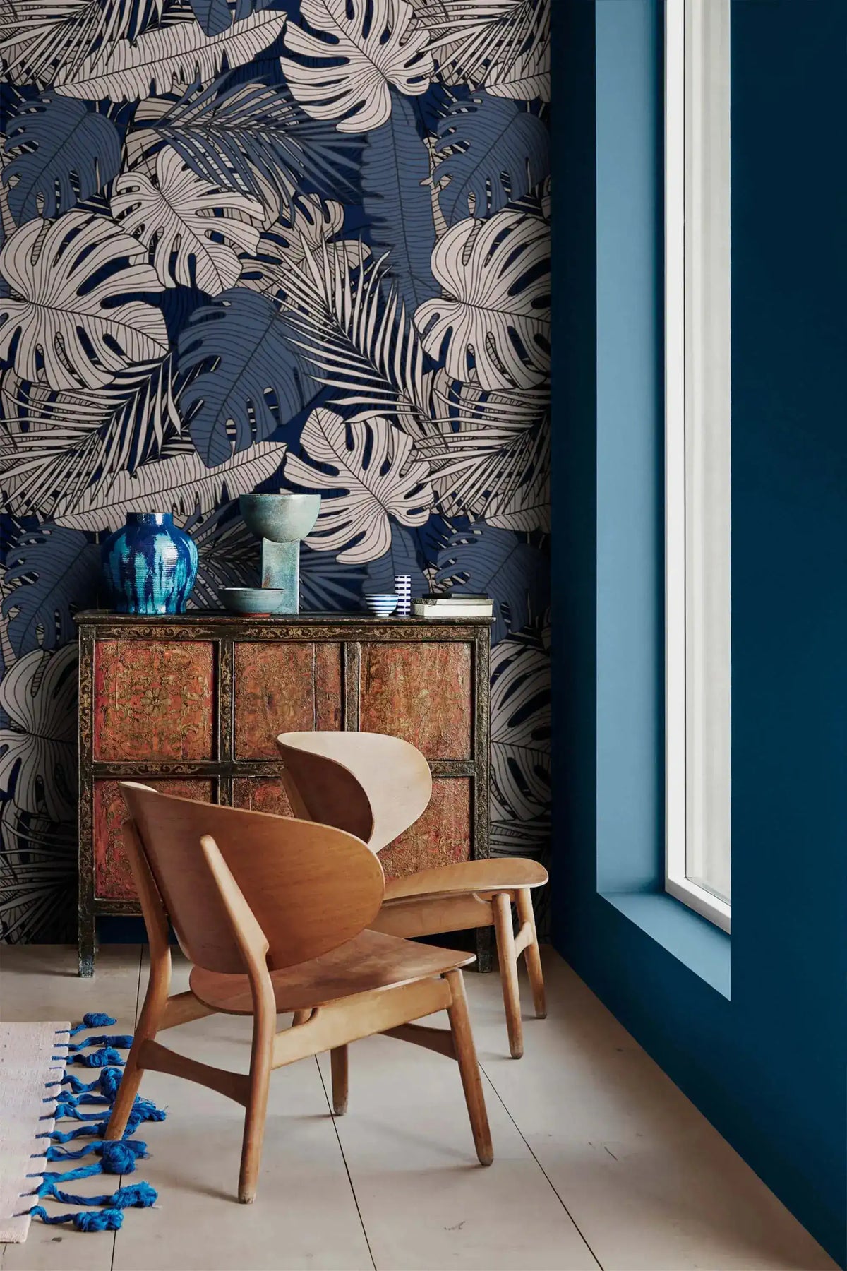 Dark navy tropical midnight leaves wallpaper with white monstera and palm fronds, in-room styling with wood armchair