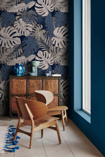 Dark navy tropical midnight leaves wallpaper with white monstera and palm fronds, in-room styling with wood armchair