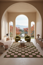 A large rectangular area rug with a dark olive green base and large dusty pink circles, styled in a modern living room with two green armchairs, a white sofa, and a view through arched windows.
