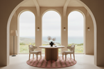 A minimalist dining area with a round wooden table and two cream upholstered chairs on a pink patterned round rug, set against a backdrop of three large arched windows overlooking a coastal landscape.