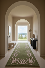 A long green area rug with a white rope-style ornamental pattern runs down a hallway framed by arched doorways, with a bench, console table, and a large window showing a lake view in the background.