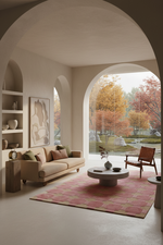 A modern living room with beige walls and large arched windows looking out onto autumn trees. The room features a beige sofa, a round coffee table, a pink patterned rug, and a wooden armchair.