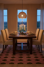 A dining room scene with a dark wood rectangular table set for six, surrounded by cream upholstered chairs. A woven pendant light hangs above, and a candelabra sits on the table.