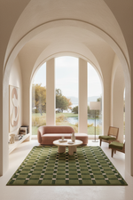 A styled interior scene with a pink sofa, green accent chair, round coffee table, and a large green patterned area rug in a bright room with arched windows and an outdoor view.