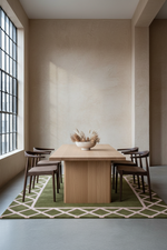 A minimalist dining room setup featuring a light wood rectangular table and four dark wood chairs arranged on a green geometric patterned rug. The room has textured beige walls, a large black-framed window on the left, and a concrete floor.