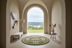 A round area rug with a botanical floral pattern in sage green and cream, styled in a modern interior setting with an arched window view of a lake and forest.