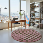 A round pink and olive green patterned area rug in a modern, sunlit loft with a wooden desk, black chairs, a large bookshelf, and a city skyline view.