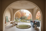 A modern living room framed by large arched openings, featuring a blue sofa, white armchair, round green rug, and a garden view outside with autumn trees.