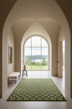 A green and beige checkered area rug laid out on a light-colored floor in a grand arched hallway with a large window overlooking a lake and green fields.