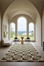 A large geometric patterned area rug in olive green and blush pink is displayed in a modern room with arched windows overlooking a lake and forest landscape. The room features a cream sofa, black chairs, and a small wooden side table.