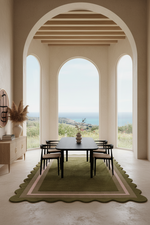 A styled interior dining room scene with a rectangular dark dining table and six matching chairs placed on a large olive green scalloped area rug. The room has warm beige walls, arched windows, and a visible coastal landscape beyond.