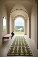 A long green and beige patterned runner rug is displayed in a grand arched hallway with a scenic lake view outside. A pink bench and a black vase with branches sit on the left side of the hallway.