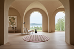 A round pink and beige patterned area rug sits on a light wood floor in a modern architectural room with arched openings and a view of water and trees.