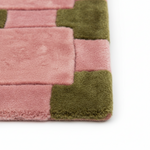 A close-up of a plush rectangular rug corner with alternating dusty pink and olive green blocks, shown on a white background.