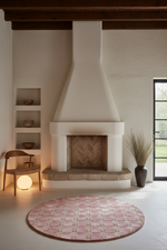 A round pink and beige woven rug in a modern minimalist living room with a white chimney stack, wooden ceiling beams, and a large window.