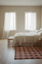 A styled bedroom interior with a bed dressed in white bedding, two tall windows with sheer white curtains, a light wood bench, and a pink and olive geometric patterned rug on a pale wood floor.