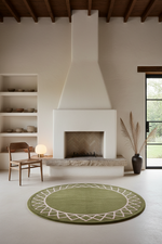A round sage green area rug with a white geometric border pattern, styled in a minimalist modern living room with a white fireplace, wooden ceiling beams, and a wooden chair.