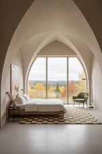 A modern bedroom with a large arched window overlooking an autumn forest landscape. The room features a low-profile bed with white bedding, a green accent chair, a patterned rug, and a high arched ceiling.