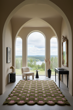 A large rectangular area rug with a scalloped edge, featuring a woven geometric pattern in olive green and dusty pink, displayed in a grand room with arched windows overlooking a lake.