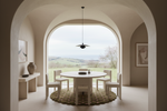 A minimalist dining room with a round white table and four matching chairs on a woven round rug, framed by a large arched opening that looks out onto a green landscape. The room has warm beige walls, a hanging pendant light, and a side console with a vase.