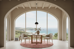 A modern dining room interior with a round wooden dining table, four upholstered chairs, a hanging pendant light, and a pink patterned round rug, all set in front of a massive arched window overlooking a coastal landscape.