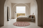 A modern minimalist bedroom with a low wooden bed, pink patterned round rug, and a large arched window showing an autumn forest view. The room features textured plaster walls and a tall black-framed mirror.