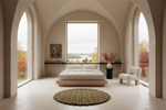 A minimalist bedroom interior with a low platform bed, white bedding, a round textured rug, a white chair, and large arched windows looking out onto a lake and autumn trees.