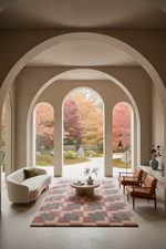 A modern living room with a large patterned area rug, a curved cream sofa, and two brown leather armchairs, set under a series of arched windows looking out onto an autumn garden with colorful foliage.