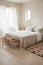 A minimalist bedroom scene featuring a neatly made bed with beige linens, white curtains, a light wood bench, and a green rug with pink leaf patterns on a light wood floor.