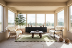 A bright, modern sunroom or conservatory with two cream-colored armchairs, a low wooden coffee table, a green patterned rug, and a black sideboard against large windows.