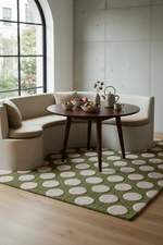 A modern living room corner featuring a curved cream banquette sofa, a round dark wood coffee table, and a green area rug with large beige polka dots. A large arched window is on the left, and a concrete wall is on the right.