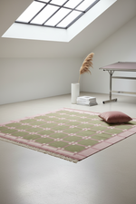 A rectangular area rug with a sage green and dusty pink geometric pattern and fringe, styled on a light gray floor in a minimalist room with a skylight, a cushion, and a vase of pampas grass.