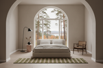 A minimalist bedroom interior with a low platform bed centered beneath a large arched window, flanked by a black floor lamp and a wooden chair. The room has warm beige walls and flooring, with a green and beige plaid rug in front of the bed.