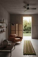 A modern living room interior with a brown leather armchair, dark wood coffee table, tall floor lamp, folding screen, and a long green runner rug with pink circular accents. An open doorway reveals greenery outside, and the room has concrete walls and ceiling.
