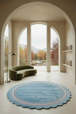 A styled interior scene with a large scalloped round blue rug in the foreground, a curved olive green sofa, two white armchairs, a small round coffee table, and tall arched windows showing an outdoor autumn landscape.