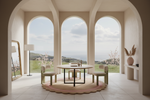 A minimalist dining set consisting of a round light wood table and four matching upholstered chairs, arranged on a scalloped pink and cream rug inside a grand room with arched windows overlooking a scenic landscape.