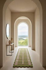 A long green runner rug with scalloped edges and pink oval motifs lies on a light floor in a narrow arched hallway leading to a large window with an outdoor landscape view.