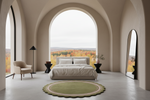 A modern bedroom interior with a large bed centered under a massive arched window overlooking an autumn forest. The room has warm beige walls, a light gray floor, a round green rug in front of the bed, a beige armchair, a black floor lamp, and a tall floor mirror.