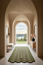 A long green patterned rug with scalloped edges lies centered in a long hallway with arched windows, showing a lake and forest view outside. A white bench, floor lamp, and wooden cabinet with a vase of branches are placed along the sides.