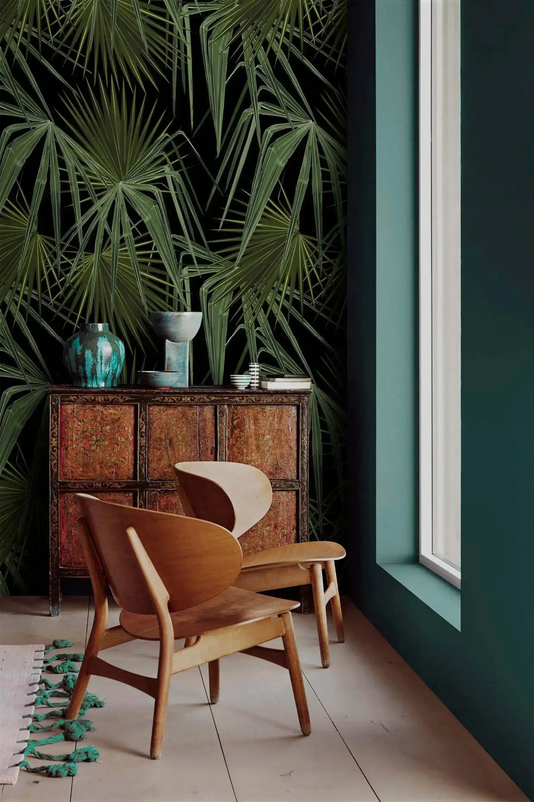 Green and black palm leaves wallpaper in corner setting with teal wall, wood Scandinavian chair and ornate antique cabinet