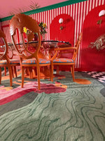 A vibrant Duo Tails Hand Tufted Area Rug adds a contemporary touch to a stylish room setting. The wool rug features an abstract design with playful colors, making it perfect for a bedroom or office. The rug is placed under a set of elegantly carved chairs, complementing the bold red and striped walls, creating a chic, modern office or bedroom space.